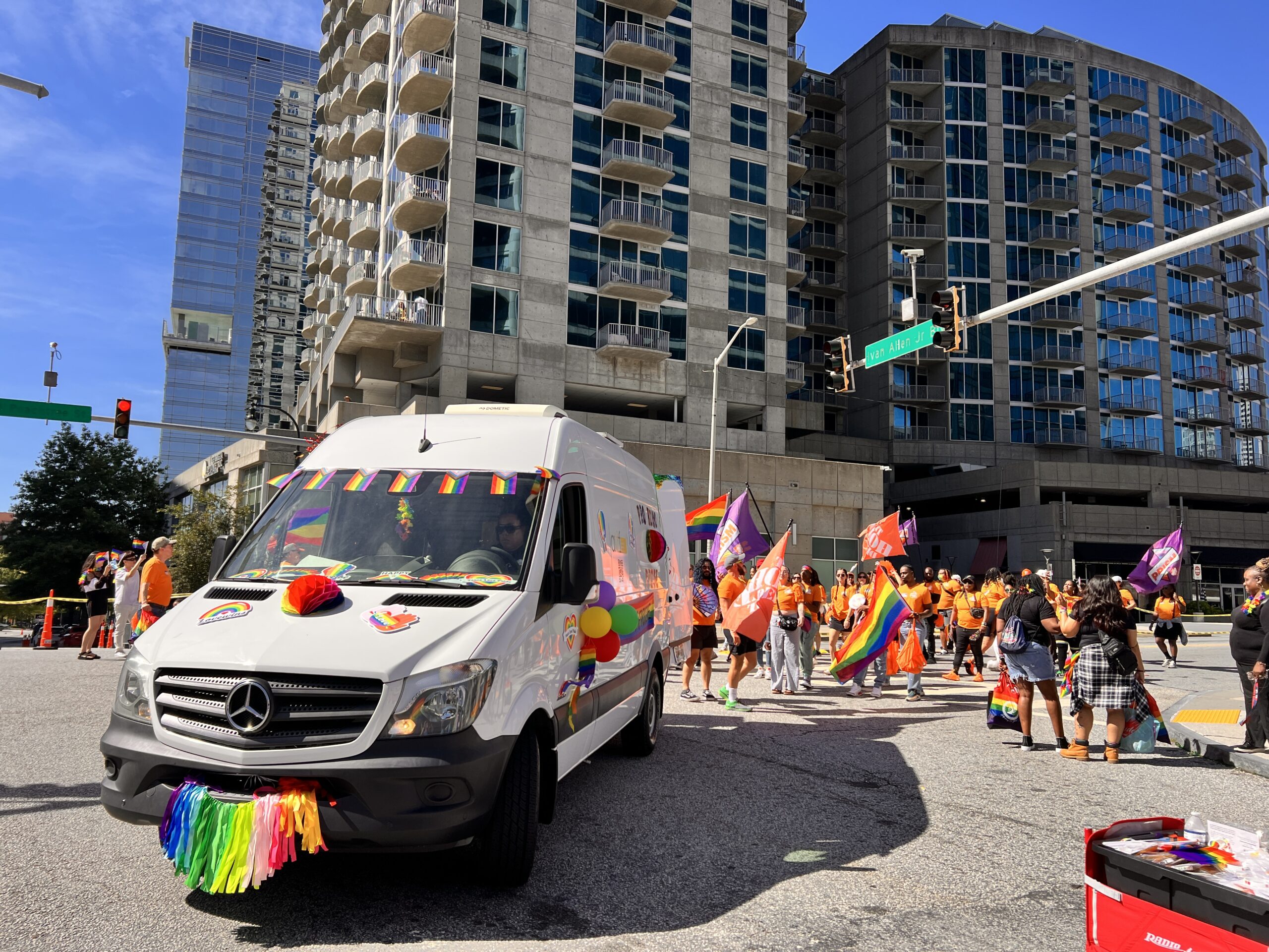 A white van decorated with rainbow pride flags and garlands leads a group of people in orange shirts and waving pride flags at a city intersection during a daytime parade. Tall buildings rise in the background.