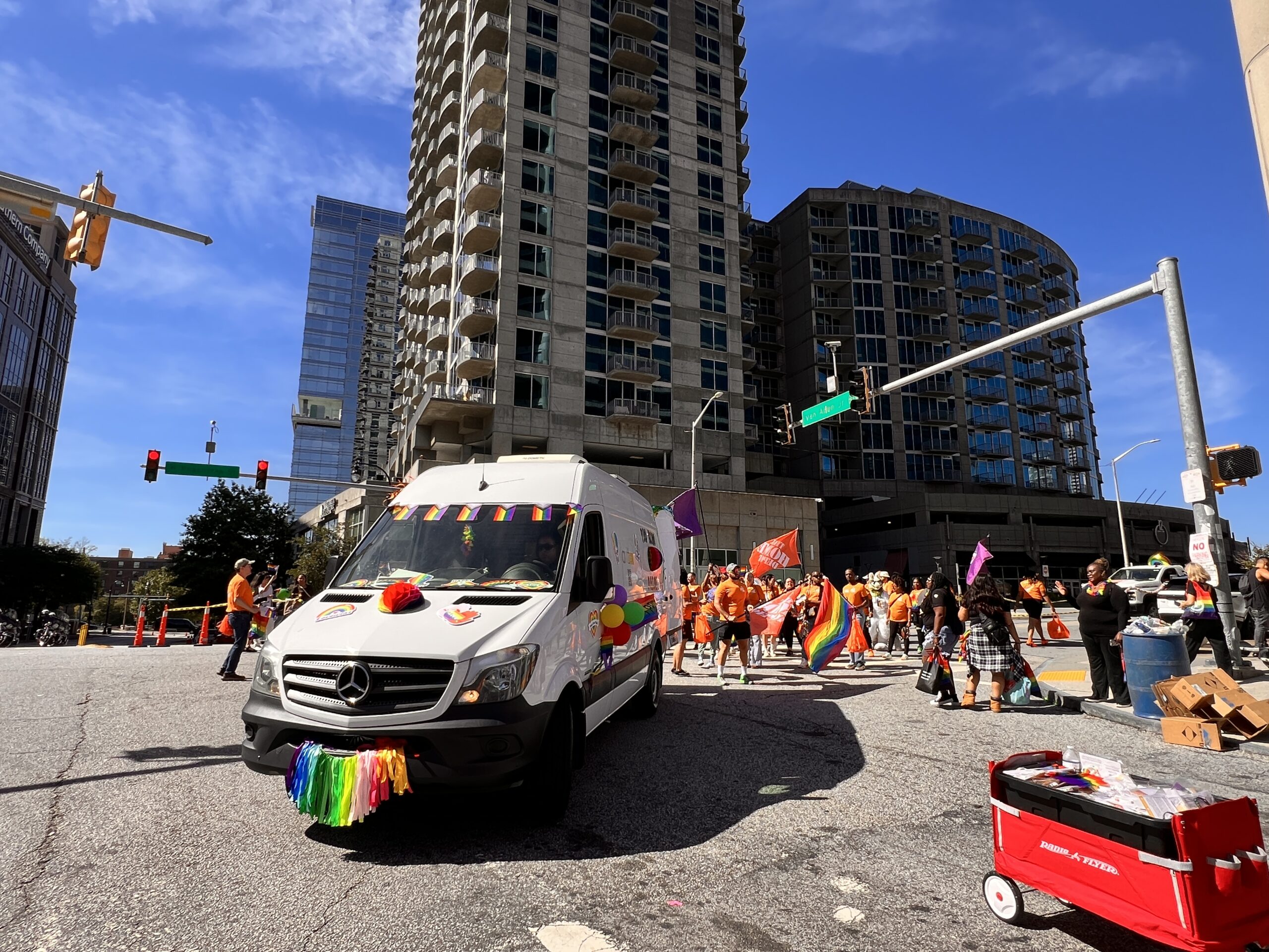 A white van decorated with rainbow flags and ribbons leads a parade on a city street, followed by a crowd of people in orange shirts holding rainbow and purple flags. Tall buildings and traffic lights are visible in the background.