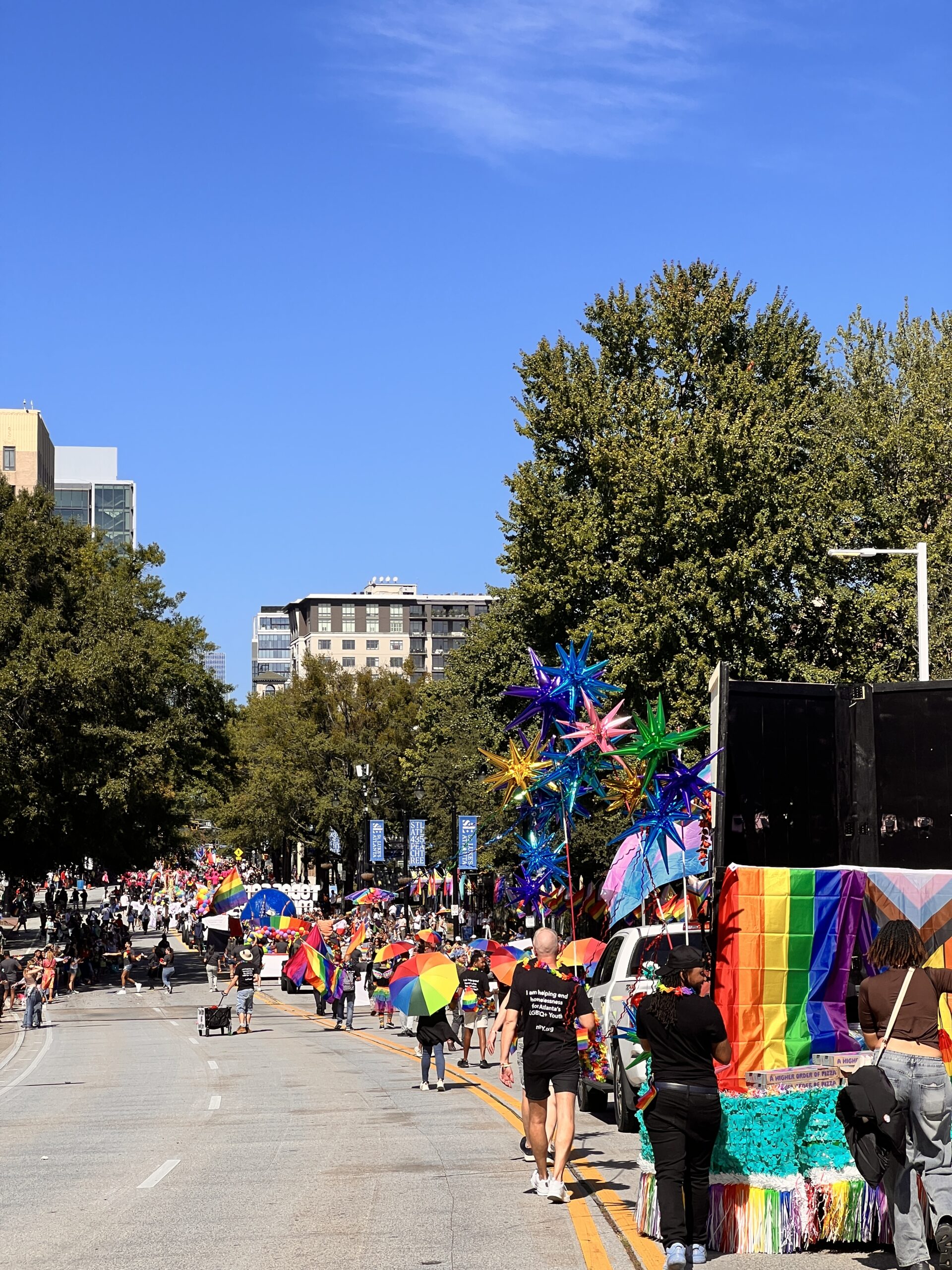 A vibrant pride parade fills a city street with people holding rainbow umbrellas, colorful star-shaped decorations, and floats draped in rainbow flags under a clear blue sky. Tall trees and city buildings line the background.