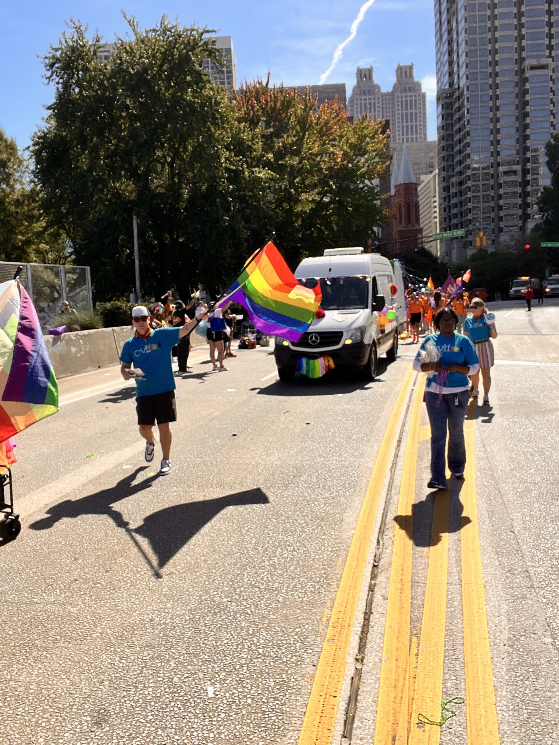 People march in a city parade holding rainbow Pride flags, with a decorated van following behind. Tall buildings and trees line the street under a sunny, clear sky.