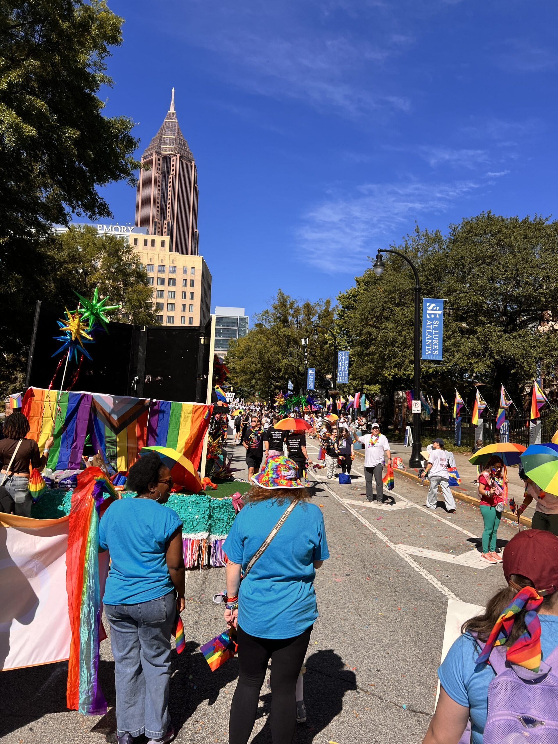 A colorful pride parade moves down a city street, with participants wearing bright clothes and rainbow flags. Tall buildings and trees line the street under a clear blue sky.