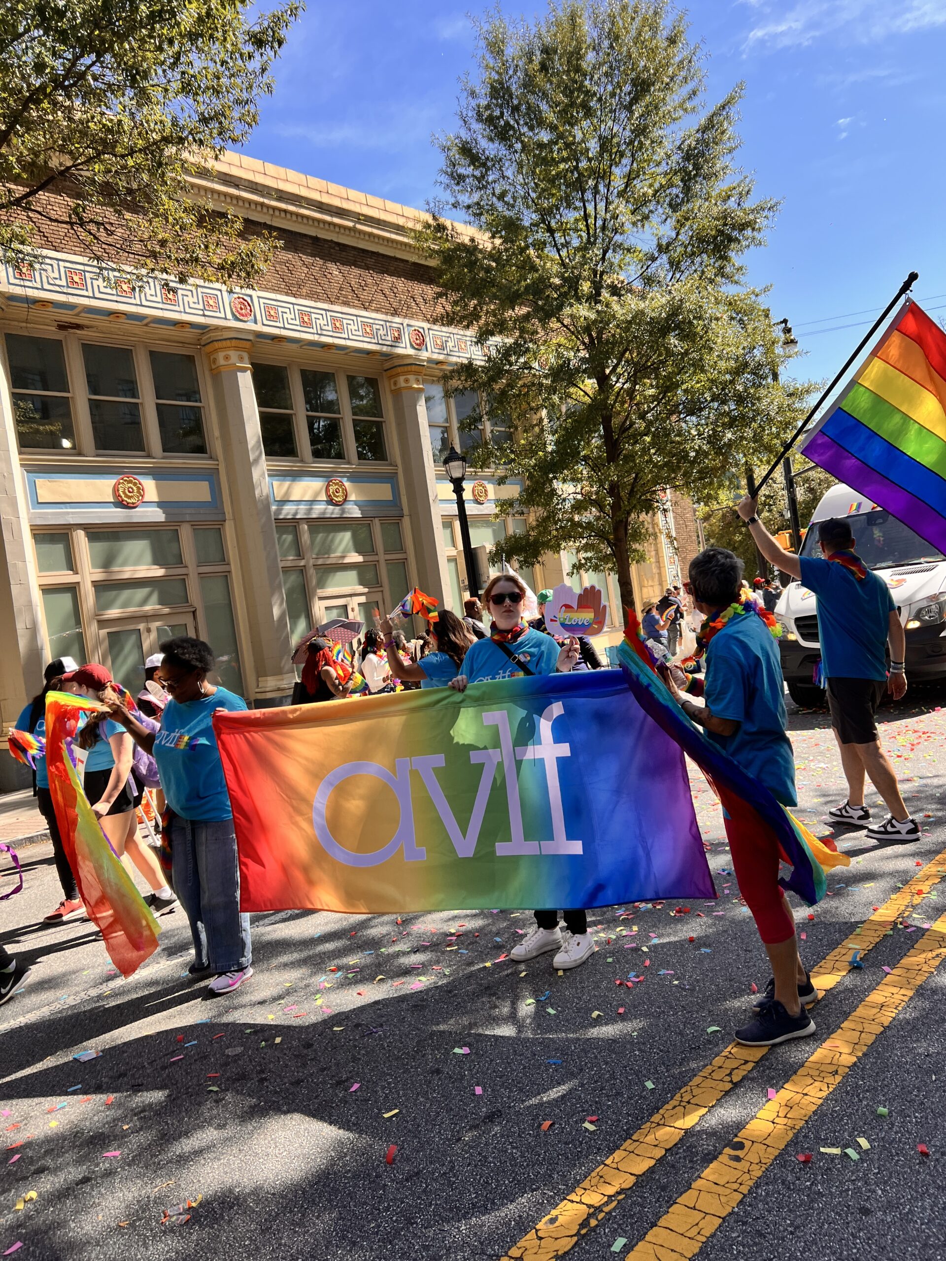 People participate in a parade, holding rainbow pride flags and a large banner with "avlf" on it. The street is lined with confetti, and the crowd is celebrating under a sunny sky in front of a decorative building.