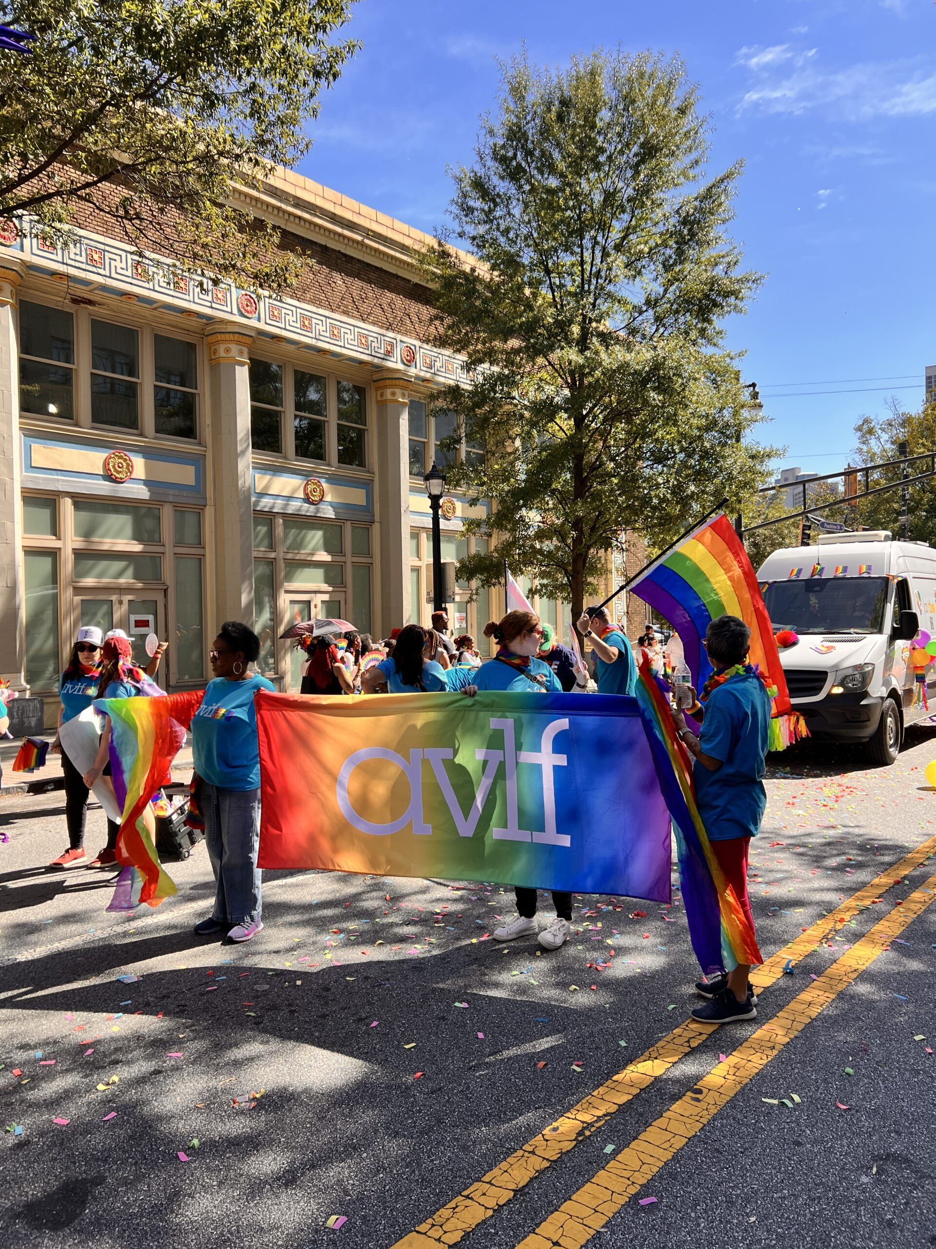 People wearing blue shirts hold a rainbow AVLF banner and pride flags at a parade on a sunny day, with a historic building and a white truck in the background. Multicolored confetti is scattered on the street.