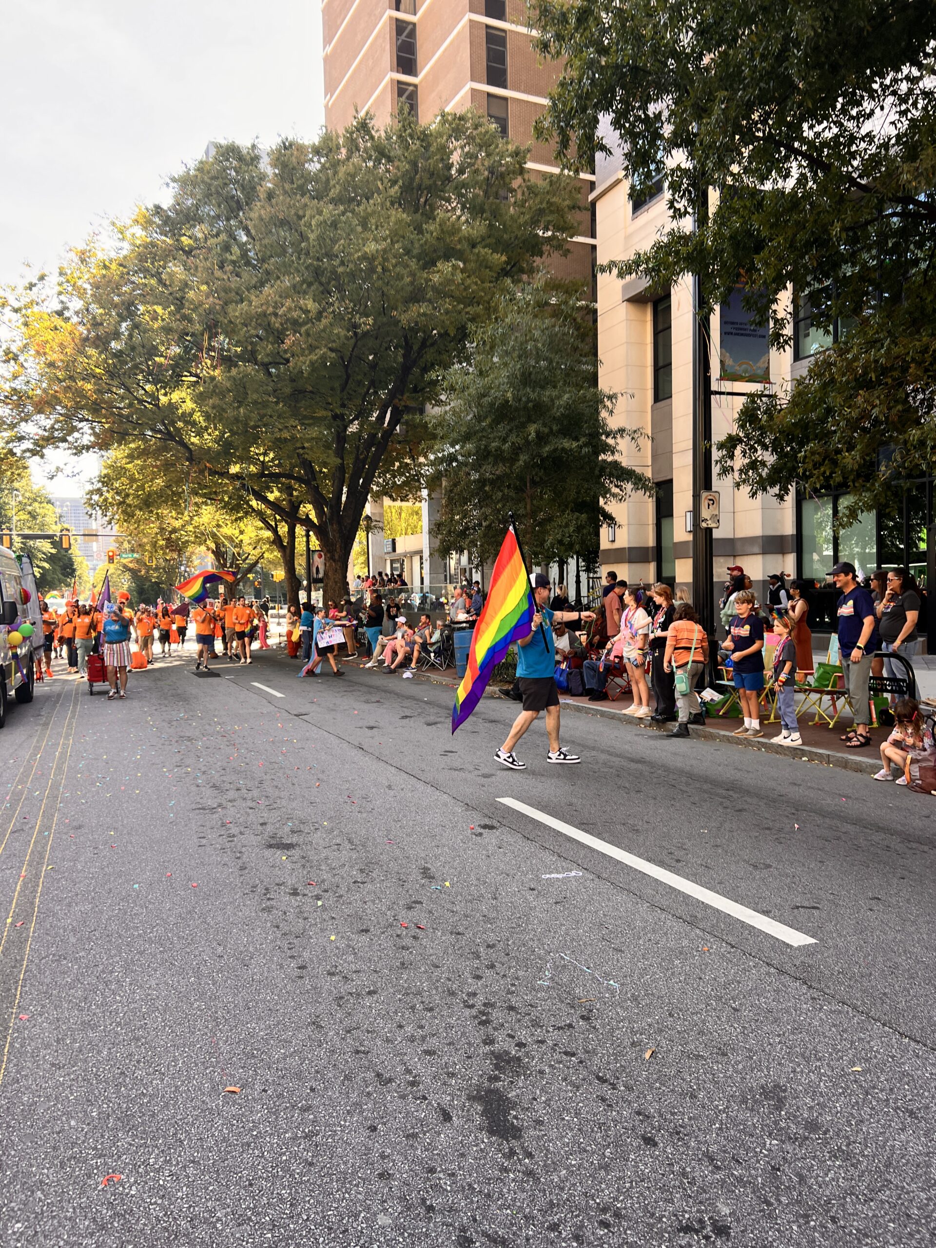 A person walks down a city street holding a large rainbow pride flag during a parade, with crowds of people watching from the sidewalk and trees lining the road.