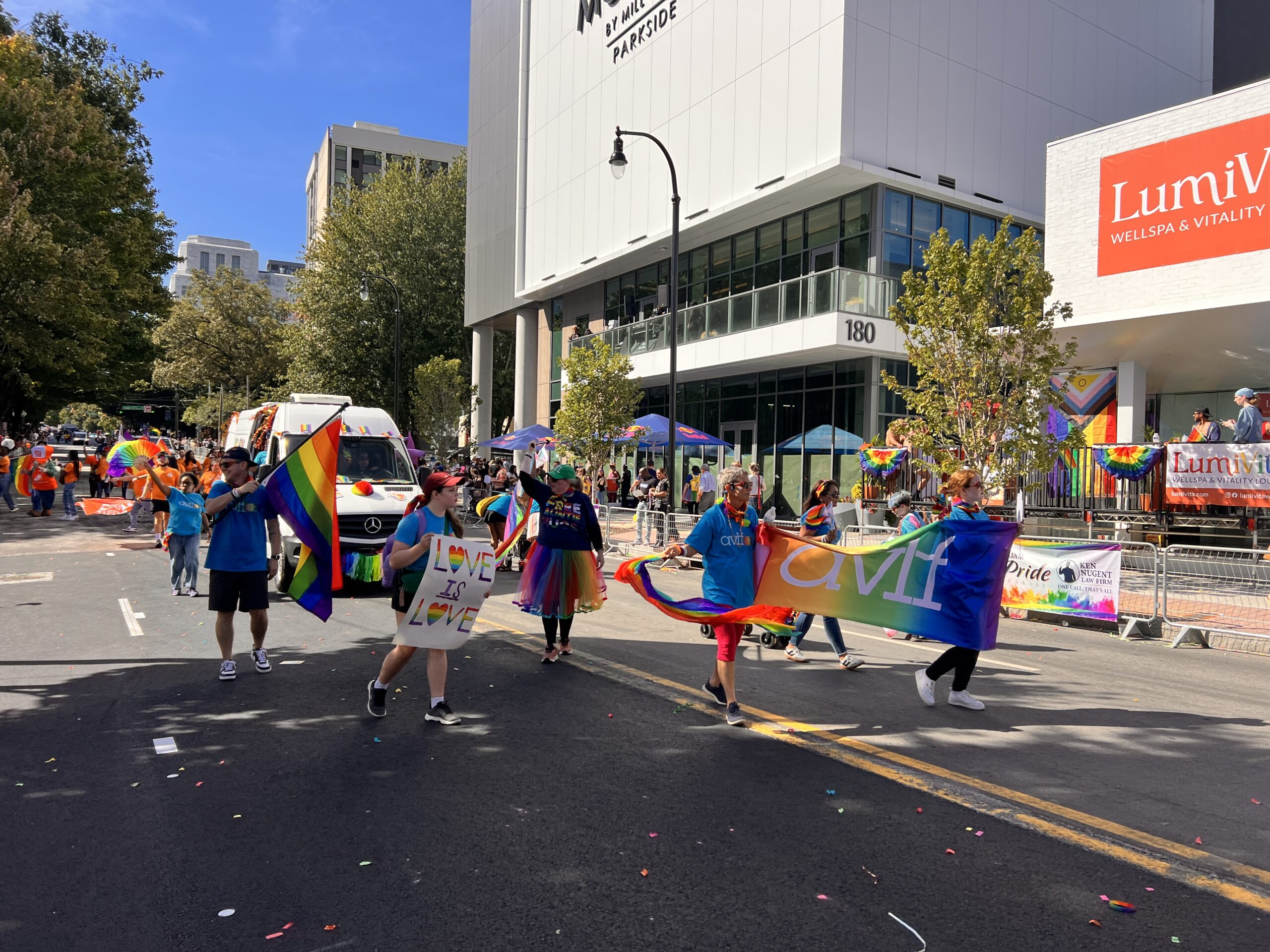 People march in a pride parade on a sunny city street, holding rainbow flags and banners. Spectators watch from sidewalks, and buildings in the background are decorated with rainbow decorations.