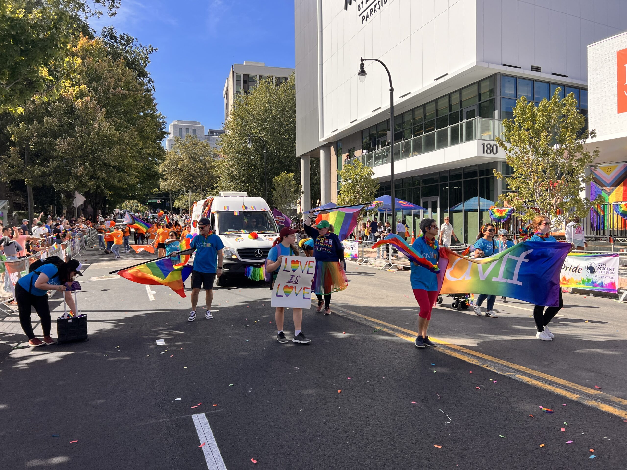 A group of people march in a pride parade, holding rainbow flags and signs that say "Love in Love." The street is lined with spectators, and there's a white van and a modern building in the background.