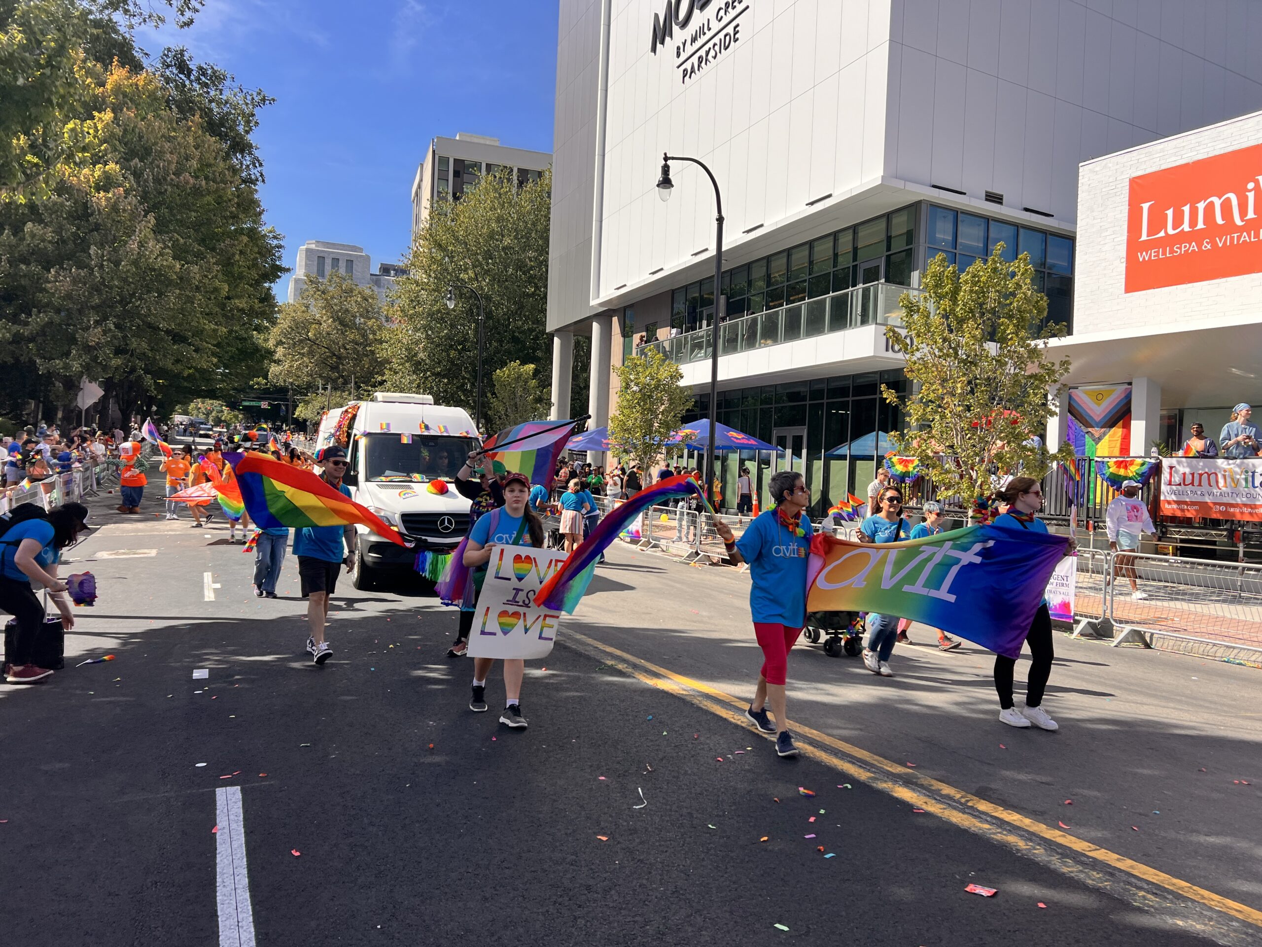 People march in a parade holding rainbow and pride flags, as well as signs reading “Love” and “Aviv.” Spectators watch from sidewalks and a building. It’s a sunny day with confetti scattered on the street.