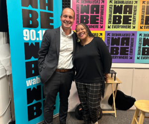 Two people smiling, standing in front of a colorful WABE 90.1 FM radio station backdrop. The man wears a suit and the woman wears a black top and plaid pants. They both appear happy and relaxed.