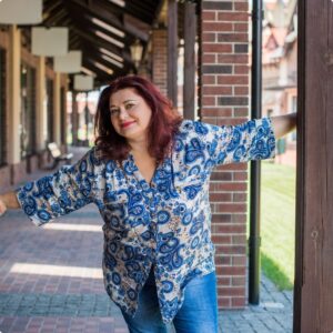 A woman with red hair, wearing a blue and white patterned blouse and jeans, smiles while standing outdoors, leaning with outstretched arms against a wooden post and brick wall. The background shows a covered walkway and grass.