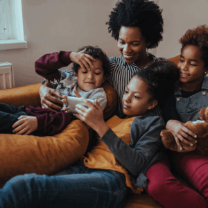 A smiling woman sits on a couch with three children. One child holds a smartphone, another leans on her lap, and the third hugs a teddy bear. They appear happy and relaxed, enjoying time together at home.