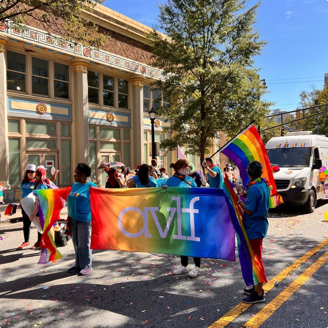 People march in a parade holding a rainbow avlf banner and rainbow flags on a sunny day. Others walk nearby, and a van decorated with balloons is in the background. A building with columns and trees line the street.