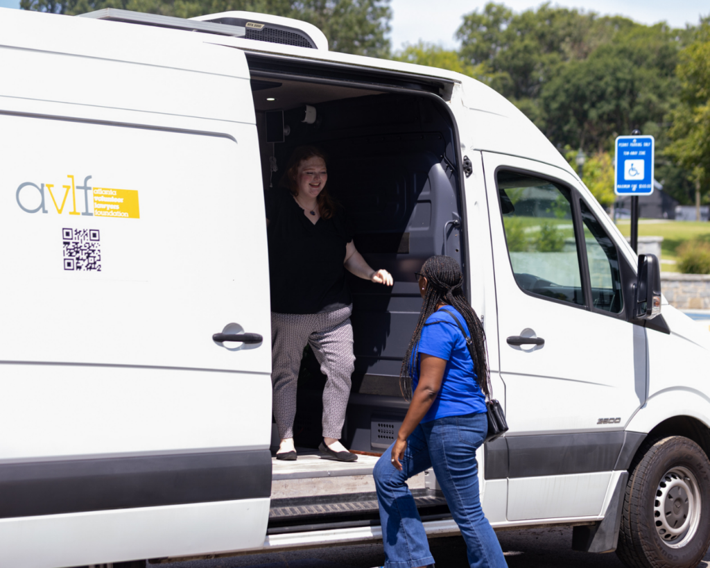 Two women interact at the open side door of a white van parked by a handicap space. One woman stands inside the van smiling, while the other stands outside looking up at her. Trees are visible in the background.