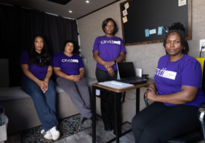 Four women wearing purple shirts sit and stand in a room with a desk, laptop, and bulletin board. They look toward the camera, appearing serious and engaged in a professional setting.