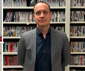 A man in a gray blazer and dark shirt stands in front of shelves filled with rows of CDs. He is facing the camera with a neutral expression.