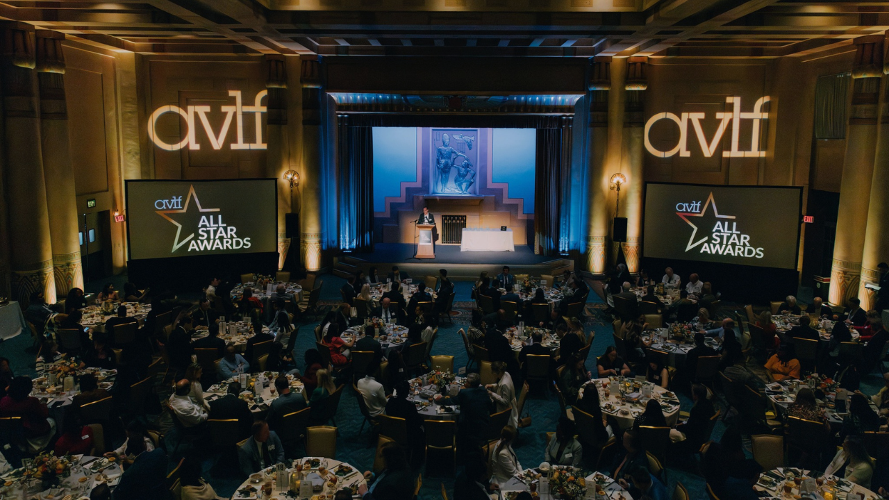 A large, elegant banquet hall hosts the AVLF All Star Awards, with round tables full of seated guests, a stage with a podium, and AVLF logos projected on the walls. Two large screens display the event name.