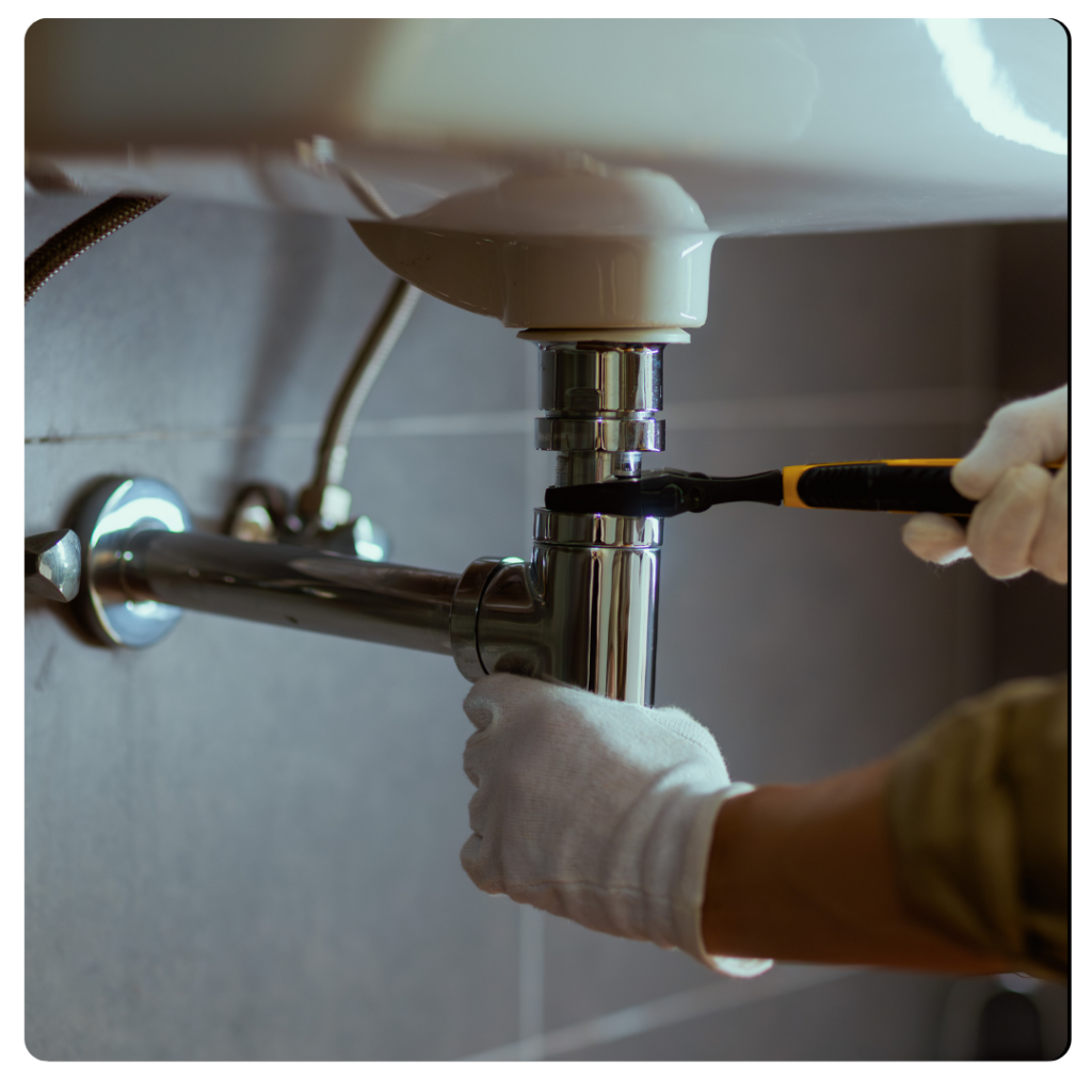 A person wearing white gloves uses a wrench to tighten or loosen a pipe under a bathroom sink against a tiled wall.
