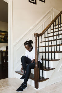 A woman in athletic wear sits on indoor stairs, tying her black sneakers. She is wearing a white top, black leggings, and sunglasses resting on her head. The setting appears to be a well-lit home interior.