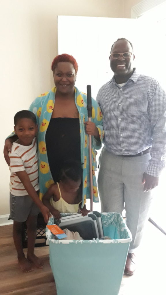 A smiling group of three people standing indoors. A woman in a duck-patterned robe holds a cane, with a young boy hugging her. A man stands next to them wearing glasses and a checkered shirt.