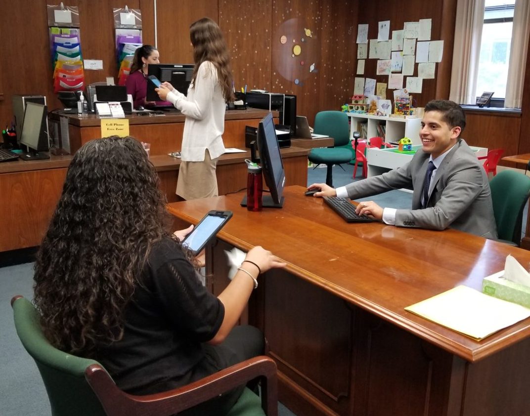 A professional office scene with a man in a suit sitting at a desk using a computer. Two women stand nearby engaged in conversation, one holding a tablet. The room has wooden walls, a bulletin board, and various colorful decorations.