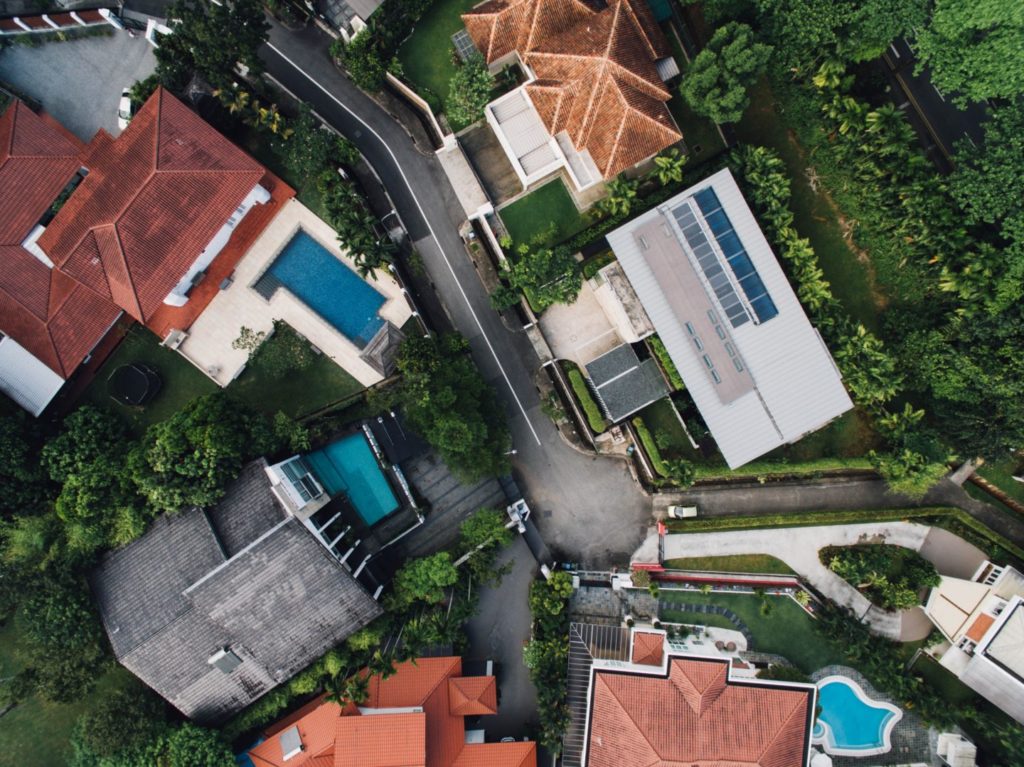Aerial view of a residential area featuring several houses with red and gray roofs, surrounded by green trees. Two houses have swimming pools. A narrow road runs through the center of the neighborhood.